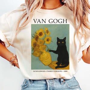 May include: A cream-coloured t-shirt displaying Van Gogh's "Sunflowers" with a black cat. The artwork shows a vase of sunflowers and the text "VAN GOGH" and "SUNFLOWERS (THIRD VERSION) - 1888". The shirt is worn with blue jeans.