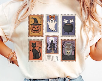 May include: Cream-colored t-shirt featuring a collection of Halloween-themed postage stamp designs. The stamps depict a jack-o'-lantern, ghost, cauldron, black cat, haunted house, and a tombstone with the words "Trick or Treat".