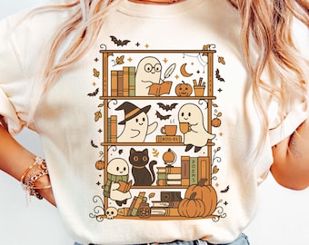 May include: Cream-colored t-shirt featuring a whimsical Halloween-themed graphic. The design showcases a bookshelf with ghosts, a black cat, pumpkins, and books. The shirt has short sleeves and a relaxed fit, perfect for casual wear.