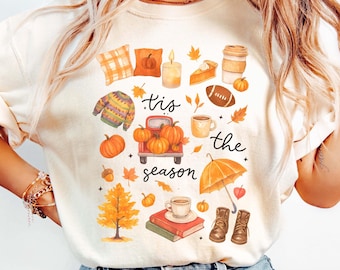 May include: Cream-colored t-shirt with a colorful fall-themed graphic. The design includes pumpkins, leaves, a truck, and the text "'tis the season". Other elements include a sweater, coffee cup, and boots.