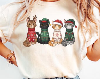 May include: Cream-colored t-shirt featuring a graphic of five cats dressed in Christmas sweaters and hats. The cats are various colors, including brown, black, orange, and gray, with festive designs like snowflakes and reindeer antlers.