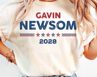 May include: Cream-colored t-shirt with the text "GAVIN NEWSOM 2028" in red and blue. The design includes a horizontal stripe with stars. The shirt is paired with light blue jeans.