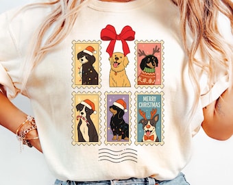 May include: Cream-colored t-shirt featuring a postage stamp design with six different dog breeds. Each dog is dressed in festive holiday attire, with a large red bow at the top. The words "MERRY CHRISTMAS" are printed on one of the stamps.