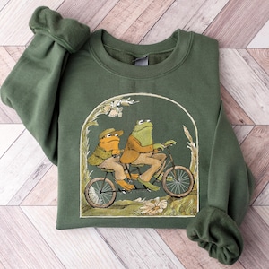 May include: Green sweatshirt with a graphic of two frogs riding a bicycle. The frogs are wearing clothes and the bicycle has a basket.