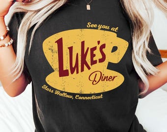 May include: Black t-shirt featuring a yellow coffee cup design with the words "Luke's Diner" in burgundy and the text "See you at" above and "Stars Hollow, Connecticut" below. The design has a distressed, vintage look.