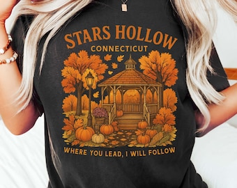 May include: Black t-shirt featuring a fall-themed graphic with a gazebo, pumpkins, and autumn foliage. The text reads "STARS HOLLOW CONNECTICUT" and "WHERE YOU LEAD, I WILL FOLLOW". The design is in shades of orange, brown, and yellow.
