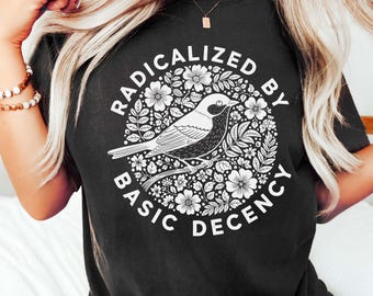 May include: Black t-shirt with a circular design featuring a bird and floral elements. The text "RADICALIZED BY BASIC DECENCY" is printed around the design in a white, sans-serif font. The shirt is a dark color and has a casual style.
