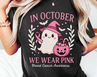 May include: Black t-shirt with a pink and white graphic design. The design includes a ghost wearing a witch hat, a pink ribbon, a jack-o'-lantern, and the text "IN OCTOBER WE WEAR PINK Breast Cancer Awareness."