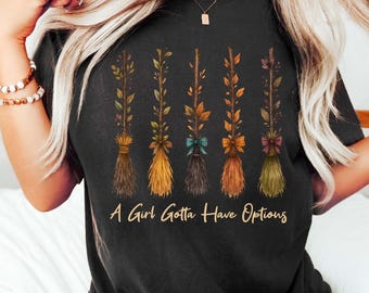 May include: Black t-shirt featuring watercolor illustrations of five decorative brooms with floral accents and bows. The text "A Girl Gotta Have Options" is printed below the brooms. The shirt is a dark color.