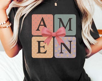 May include: Black t-shirt with the word "AMEN" spelled out in colorful blocks. The blocks are decorated with patterns and a pink bow. The shirt is a comfortable, casual style.