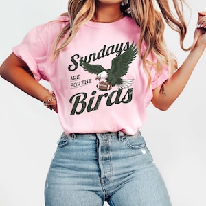 May include: Pink t-shirt featuring the text "Sundays are for the Birds" and an eagle graphic holding a football. The eagle has dark green wings and a white head. The shirt is worn with blue jeans.