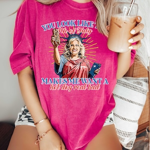 May include: A bright pink t-shirt with a graphic of the Statue of Liberty holding a hot dog. The text on the shirt reads "You look like the 4th of July" and "Makes me want a hot dog real bad."