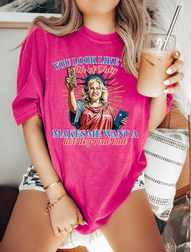 May include: A bright pink t-shirt with a graphic of the Statue of Liberty holding a hot dog. The text on the shirt reads "You look like the 4th of July" and "Makes me want a hot dog real bad."