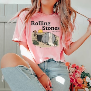 May include: A light pink t-shirt featuring the text "Rolling Stones Since AD 33" above an illustration of an open tomb with a cross and a setting sun. The shirt is a solid color and has short sleeves.
