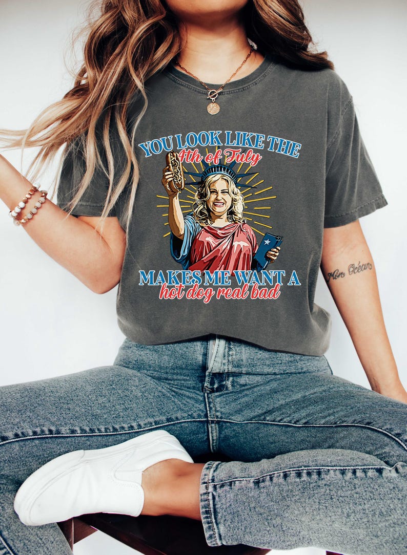 May include: Dark gray t-shirt with a graphic of the Statue of Liberty holding a hot dog and a small book. The text on the shirt reads "YOU LOOK LIKE THE 4th of July MAKES ME WANT A hot dog real bad."