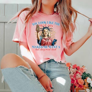 May include: A pink t-shirt with the text "YOU LOOK LIKE THE 4TH OF JULY MAKES ME WANT A hot dog real bad." The graphic features a cartoon of the Statue of Liberty holding a hot dog and a phone.