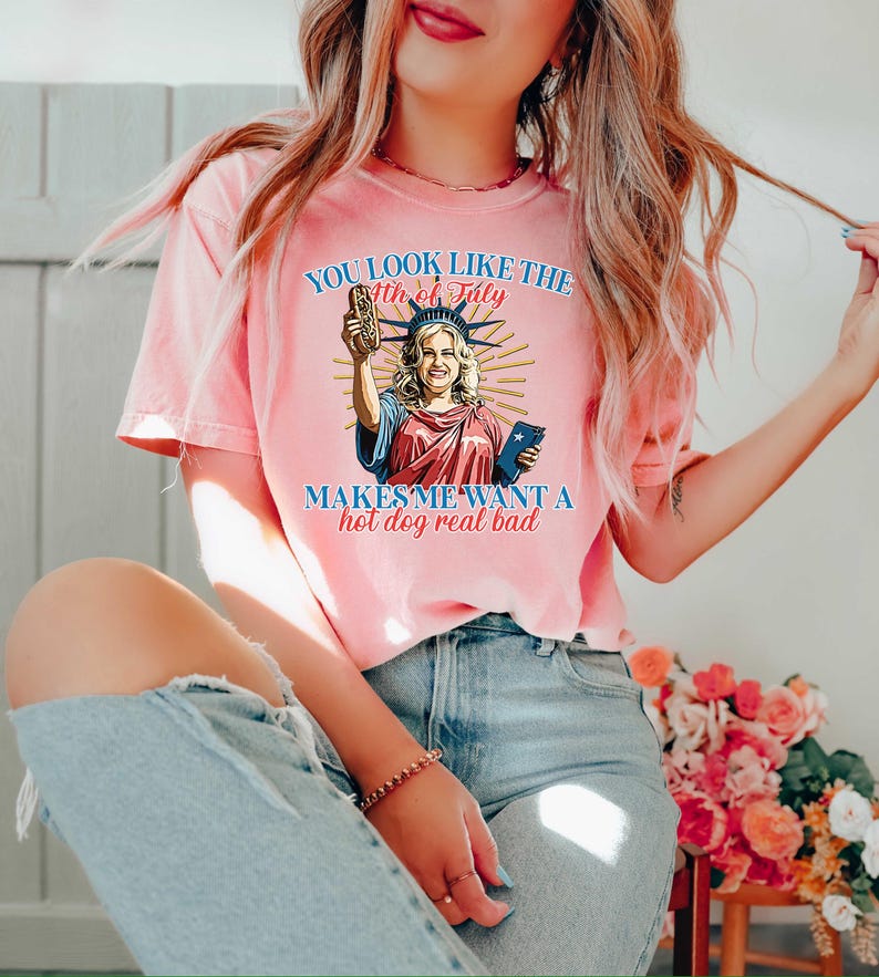 May include: A pink t-shirt with the text "YOU LOOK LIKE THE 4TH OF JULY MAKES ME WANT A hot dog real bad." The graphic features a cartoon of the Statue of Liberty holding a hot dog and a phone.