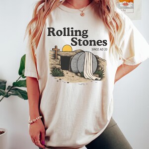 May include: Cream-colored t-shirt with the text "Rolling Stones" above an illustration of an open tomb with a cross and a rising sun. The words "SINCE AD 33" are also printed on the shirt. The shirt is worn by a person.