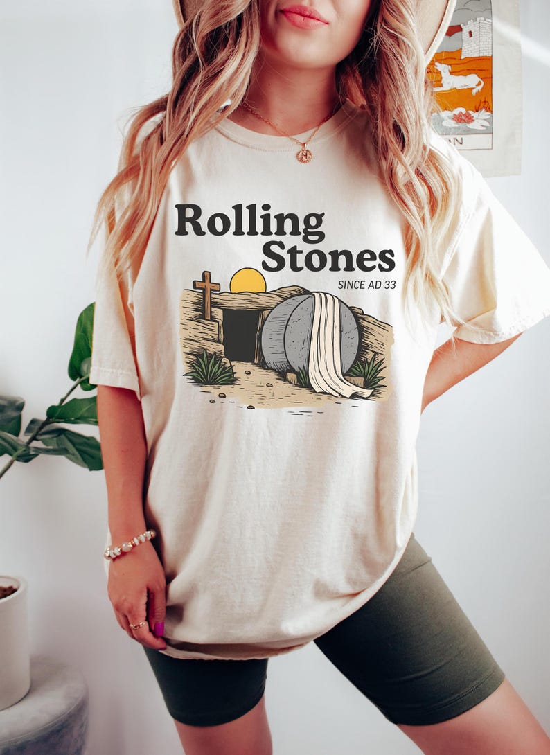 May include: Cream-colored t-shirt with the text "Rolling Stones" above an illustration of an open tomb with a cross and a rising sun. The words "SINCE AD 33" are also printed on the shirt. The shirt is worn by a person.