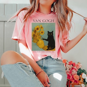 May include: A light pink t-shirt featuring a print of Van Gogh's "Sunflowers" with a black cat. The text "VAN GOGH" is above the image, and "SUNFLOWERS (THIRD VERSION) - 1888" is below. The shirt is a casual, short-sleeved style.