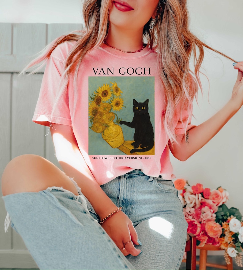 May include: A light pink t-shirt featuring a print of Van Gogh's "Sunflowers" with a black cat. The text "VAN GOGH" is above the image, and "SUNFLOWERS (THIRD VERSION) - 1888" is below. The shirt is a casual, short-sleeved style.