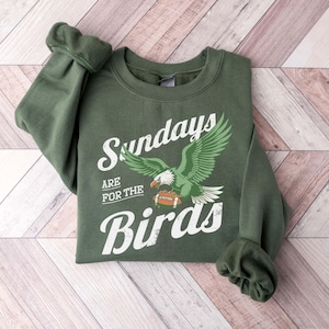May include: An olive green sweatshirt with the text "Sundays are for the Birds" in white script. A green and white eagle graphic is centered below the text, holding a football. The sweatshirt is folded on a light wood surface.