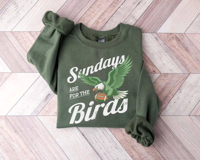 May include: An olive green sweatshirt with the text "Sundays are for the Birds" in white script. A green and white eagle graphic is centered below the text, holding a football. The sweatshirt is folded on a light wood surface.