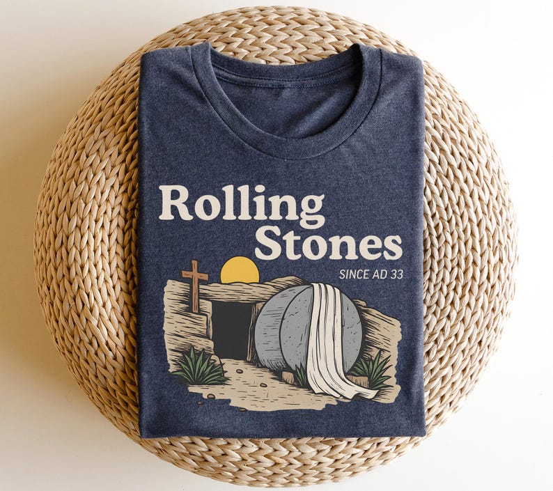May include: Navy blue t-shirt with the text "Rolling Stones" and an illustration of a tomb with a cross, sun, and stone. The text "SINCE AD 33" is also included in the design.