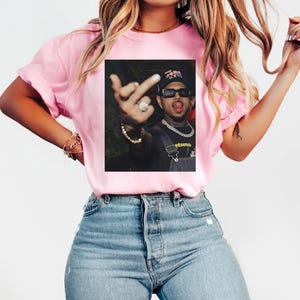 May include: Pink t-shirt with a printed image of a person wearing sunglasses, a hat, and a chain. The person is sticking out their tongue and giving the middle finger. The shirt is being worn by a person wearing blue jeans.