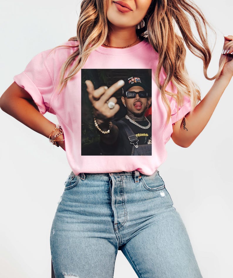May include: Pink t-shirt with a printed image of a person wearing sunglasses, a hat, and a chain. The person is sticking out their tongue and giving the middle finger. The shirt is being worn by a person wearing blue jeans.