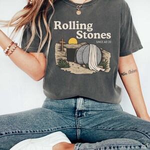 May include: Dark gray t-shirt with the text "Rolling Stones SINCE AD 33" above an illustration of a tomb with a cross, a rising sun, and a large stone. The shirt is paired with blue jeans and white sneakers.