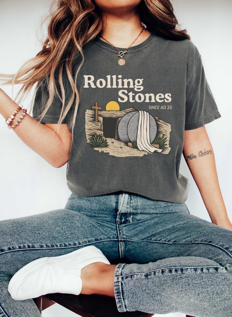 May include: Dark gray t-shirt with the text "Rolling Stones SINCE AD 33" above an illustration of a tomb with a cross, a rising sun, and a large stone. The shirt is paired with blue jeans and white sneakers.