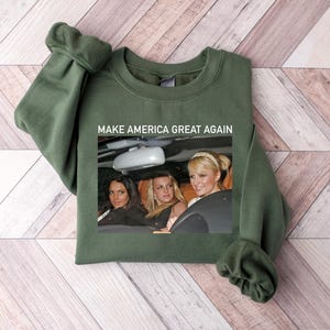 Puede incluir: Una sudadera verde oscuro con el texto "Make America Great Again" impreso en la parte delantera. La sudadera presenta una foto de tres mujeres en un coche.