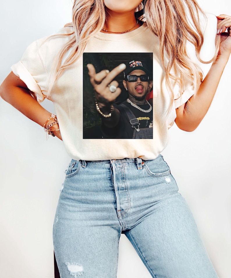 May include: Cream-colored t-shirt featuring a framed image of a person wearing sunglasses, a hat, and a chain. The person is sticking out their tongue and giving the middle finger. The shirt is paired with light blue jeans.