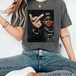 May include: Dark gray t-shirt with a framed image of a person wearing sunglasses, a hat, and a chain. The person is sticking out their tongue and giving the middle finger. The shirt is paired with blue jeans and white sneakers.