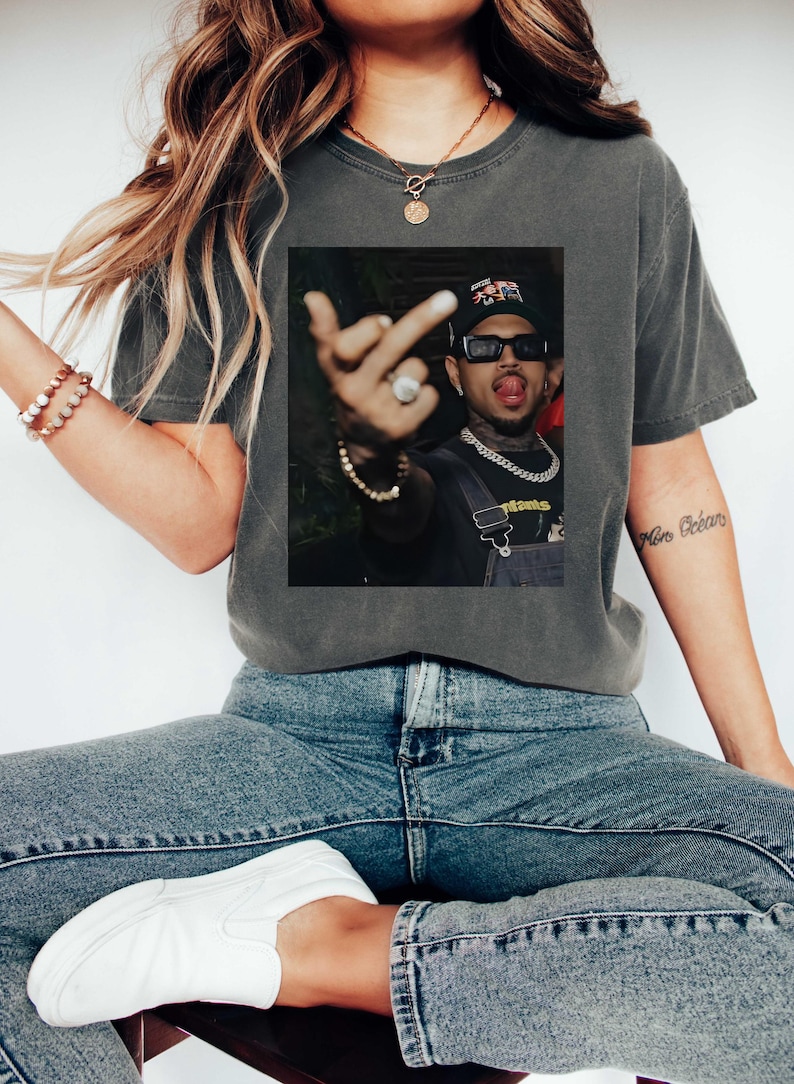 May include: Dark gray t-shirt with a framed image of a person wearing sunglasses, a hat, and a chain. The person is sticking out their tongue and giving the middle finger. The shirt is paired with blue jeans and white sneakers.