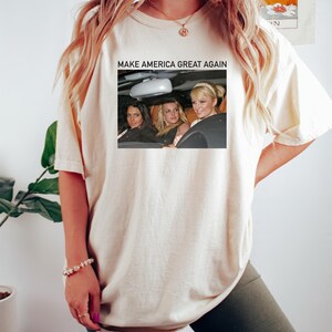 Puede incluir: Una camiseta blanca con el texto "Make America Great Again" impreso en la parte delantera. El texto est&aacute; en negro y est&aacute; centrado en la camiseta. Debajo del texto hay una foto de tres mujeres en un coche. Las mujeres llevan gafas de sol y sonr&iacute;en. La foto est&aacute; en color y est&aacute; rodeada por un borde negro.