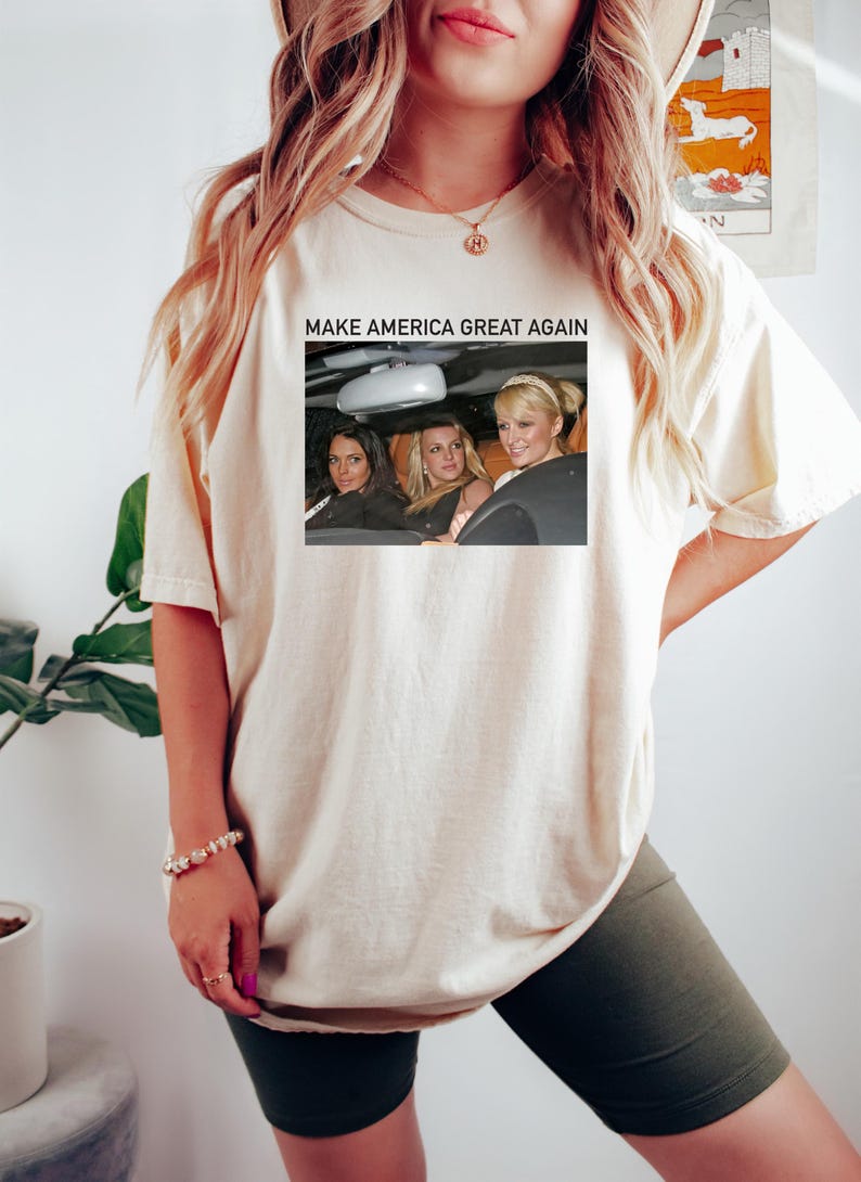 Puede incluir: Una camiseta blanca con el texto "Make America Great Again" impreso en la parte delantera. El texto est&aacute; en negro y est&aacute; centrado en la camiseta. Debajo del texto hay una foto de tres mujeres en un coche. Las mujeres llevan gafas de sol y sonr&iacute;en. La foto est&aacute; en color y est&aacute; rodeada por un borde negro.