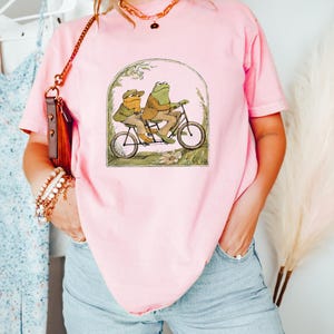 Może przedstawiać: R&oacute;żowy t-shirt z grafiką przedstawiającą dwie żaby jadące na rowerze tandemowym. Żaby mają na sobie czapki i są otoczone kwiatami.