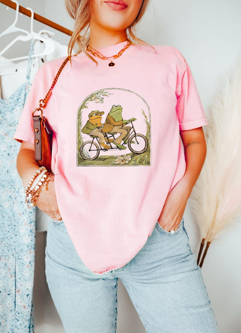 Może przedstawiać: R&oacute;żowy t-shirt z grafiką przedstawiającą dwie żaby jadące na rowerze tandemowym. Żaby mają na sobie czapki i są otoczone kwiatami.