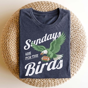 May include: A navy blue t-shirt with the text "Sundays are for the Birds" in white script. The shirt features a green and white eagle graphic holding a football. The shirt is folded on a woven, circular surface.