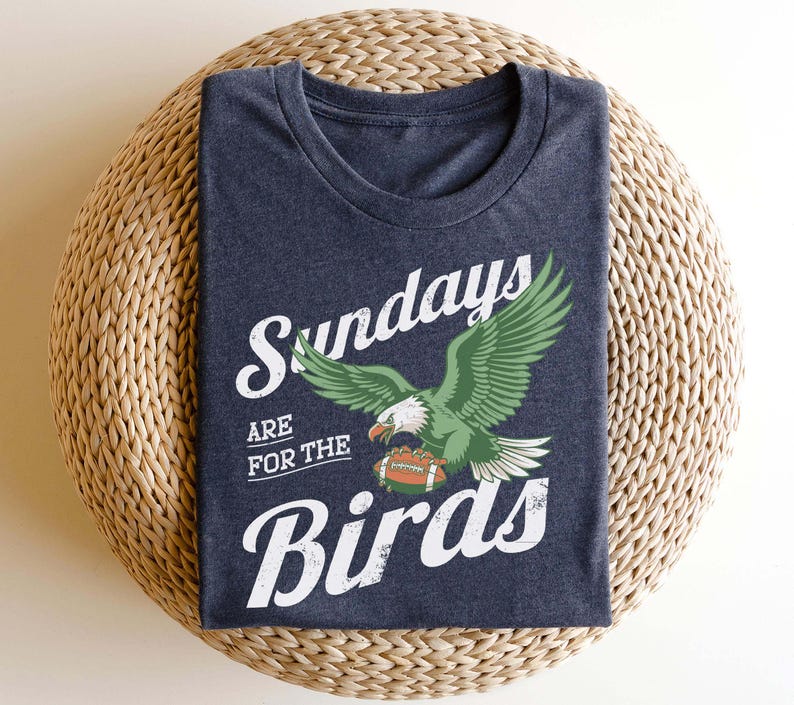 May include: A navy blue t-shirt with the text "Sundays are for the Birds" in white script. The shirt features a green and white eagle graphic holding a football. The shirt is folded on a woven, circular surface.