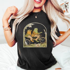 Może przedstawiać: Czarny T-shirt z grafiką przedstawiającą dwie żaby jadące na rowerze w zielonym i brązowym krajobrazie. Żaby mają na sobie czapki i kurtki.