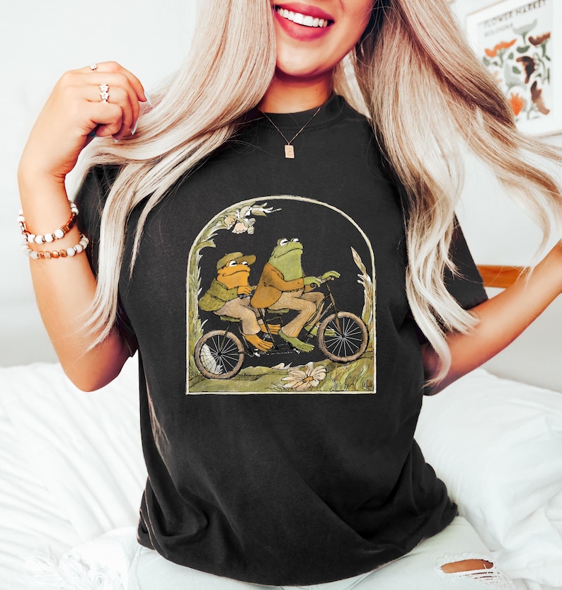 Może przedstawiać: Czarny T-shirt z grafiką przedstawiającą dwie żaby jadące na rowerze w zielonym i brązowym krajobrazie. Żaby mają na sobie czapki i kurtki.