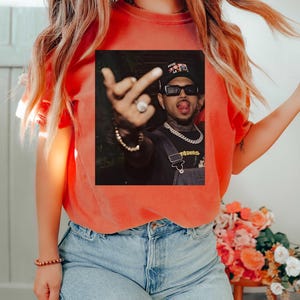 May include: Orange t-shirt with a graphic of a person wearing sunglasses, a hat, and a chain. The person is sticking out their tongue and giving the middle finger. The shirt is paired with blue jeans.