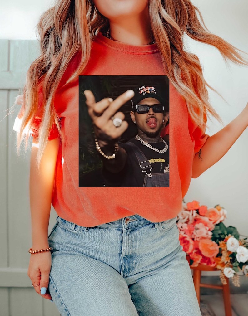 May include: Orange t-shirt with a graphic of a person wearing sunglasses, a hat, and a chain. The person is sticking out their tongue and giving the middle finger. The shirt is paired with blue jeans.