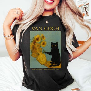 May include: Black t-shirt featuring a print of Van Gogh's "Sunflowers" with a black cat. The design includes the text "VAN GOGH" above the image and "SUNFLOWERS (THIRD VERSION) - 1888" below. The artwork depicts sunflowers in a vase.