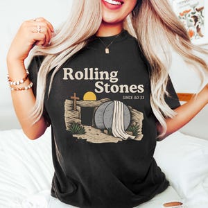 May include: Black t-shirt with the text "Rolling Stones" above an illustration of an empty tomb with a cross, sun, and draped cloth. The text "SINCE AD 33" is below the main text.