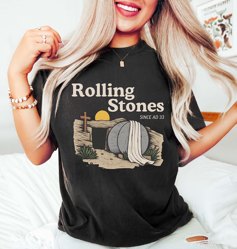May include: Black t-shirt with the text "Rolling Stones" above an illustration of an empty tomb with a cross, sun, and draped cloth. The text "SINCE AD 33" is below the main text.
