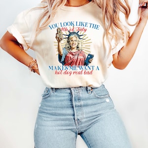May include: Cream-colored t-shirt with a graphic of a woman dressed as the Statue of Liberty holding a hot dog. The text on the shirt reads "YOU LOOK LIKE THE 4th of July MAKES ME WANT A hot dog real bad."